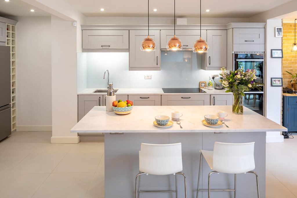A view across a bright open plan kitchen space in a Mews House with white units and a set of brass light fittings.