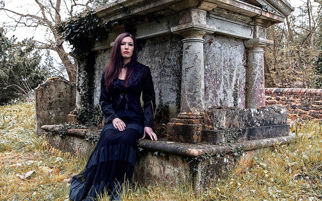 A young lady dressed in a balck victorian gothic costume sits at a mausoleum in a cemetry