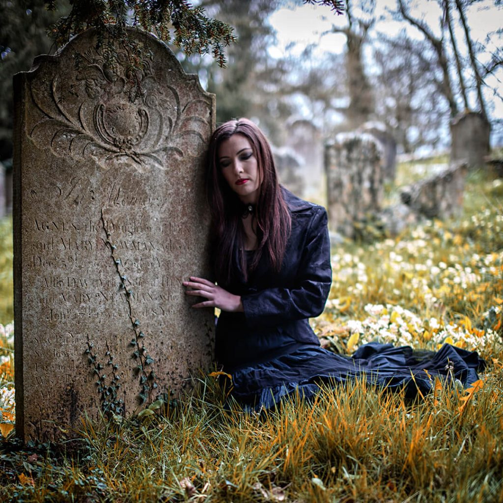 A young lady full of thought is sitting with her hand touching a gravestone in a gothic style black dress.