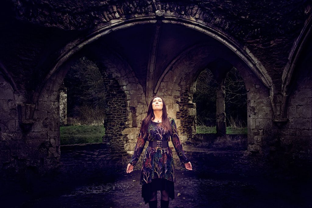 A woman is highlighted gazing skywards at a ruined cistercian abbey in a period costume which could be worn by a witch.