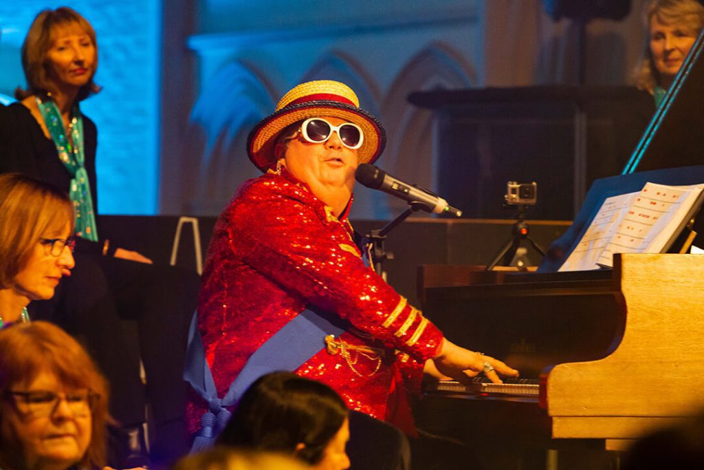 An EltonJohn tribute act is sitting at his piano singing at an event organised by th eElmbridge Ladies Choir