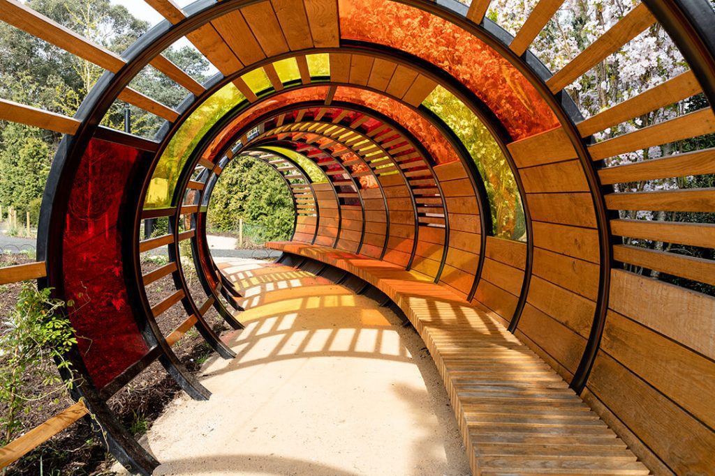 A curved route through a tunnel of coloured glass with a curved bench along the side.