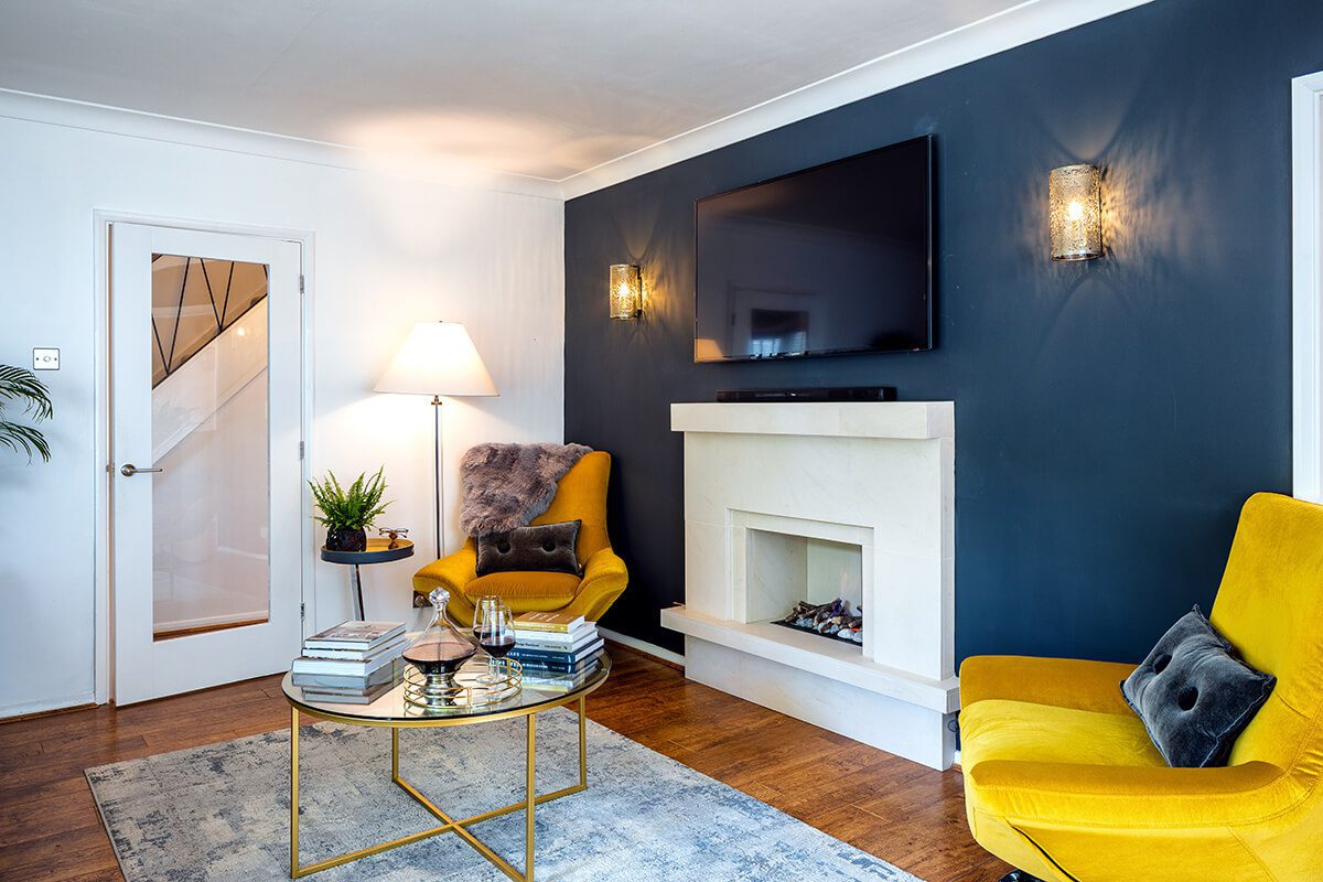 Corner of a room with a dark blue feature wall and contemporary white fireplace. There are two bright yellow easy chairs adjacent to the fireplace.