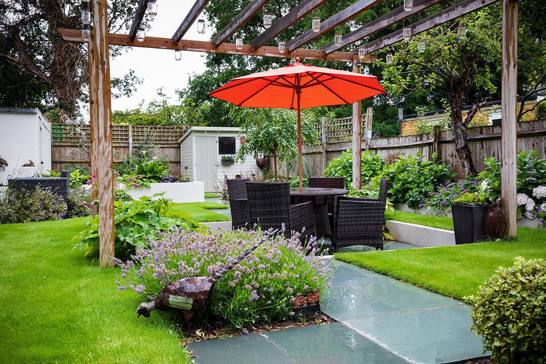 A view of a sunken area of a garden design used for entertaining. There is a pergola over rattan furniture and a garden umbrella