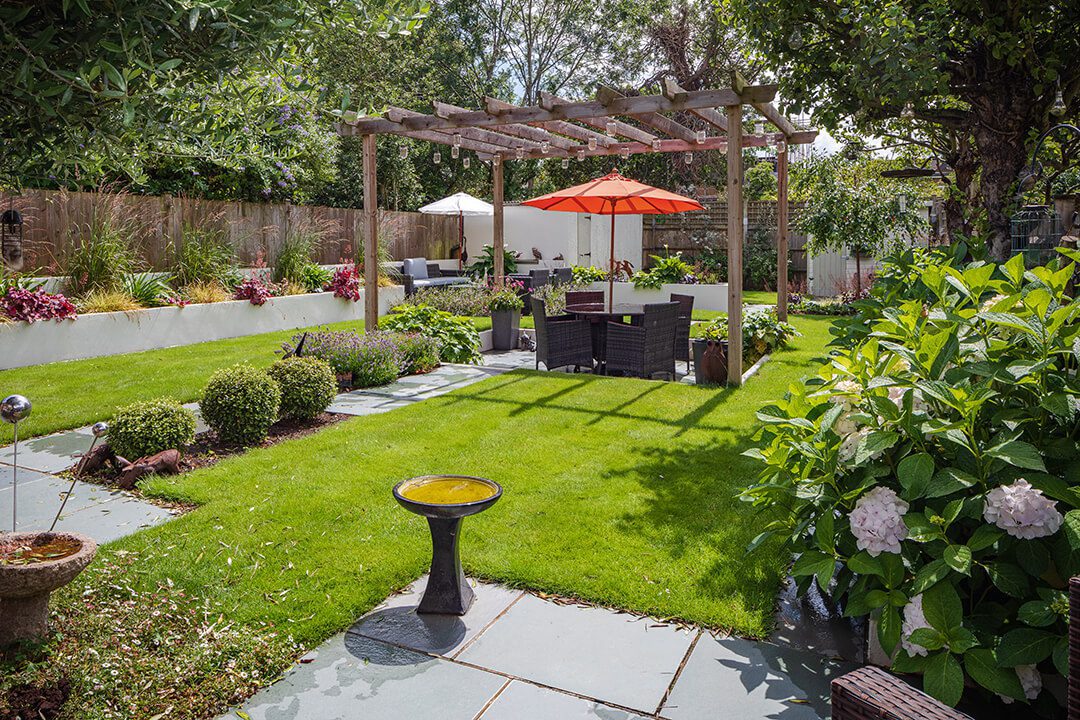 A view across a garden design featuring a sunken seating area covered by a wooden pergola with rattan seating and a sunshade.